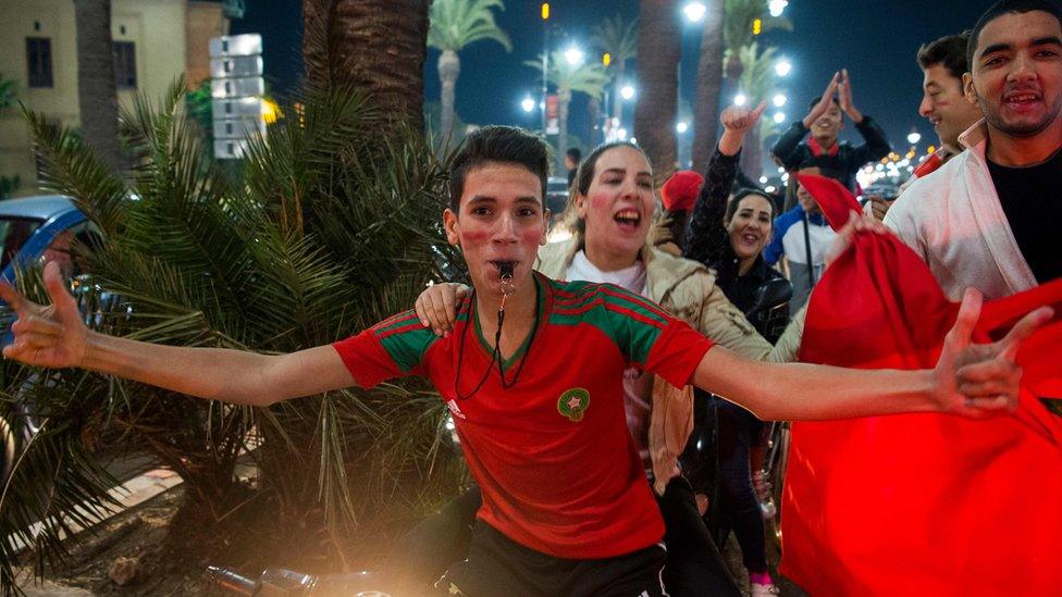 Boy wearing shirt in the Moroccan colours red and green blows a whistle as people behind him dance