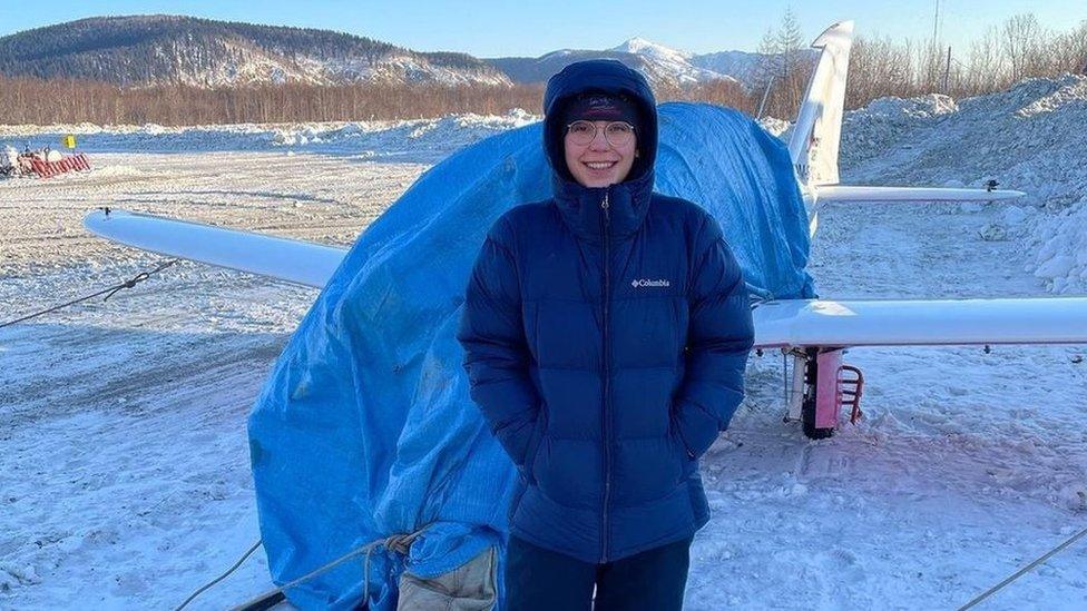 Zara Rutherford in Ayan standing in the snow in front of her aircraft which is covered in a blue tarpaulin