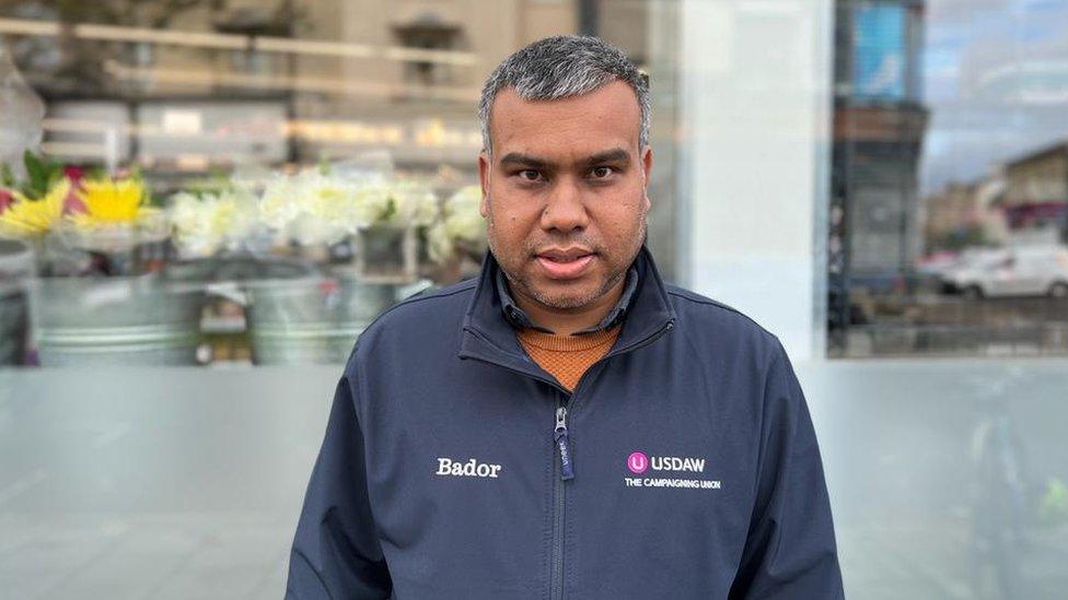 Bador is standing outside a supermarket window. In the background you can see buckets of flowers. He is wearing a blue fleece which has his name on the right hand side and a logo, for USDAW the shop workers union.