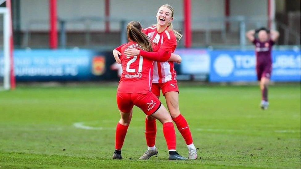 Daisy Clements celebrating a goal for Stourbridge FC, wearing a red kit with white "21" visible on the back of the shirt. There is green grass and a blurred background.