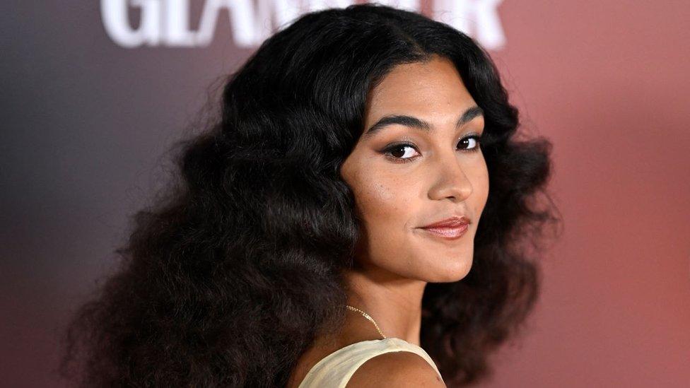 Olivia Dean - a 24-year-old woman with long dark curly hair worn loose down her back. The singer has brown eyes and smiles at the camera over her right shoulder. She is pictured at a red carpet event and wears a short sleeved cream dress, winged black eyeliner and a rose coloured lip gloss.