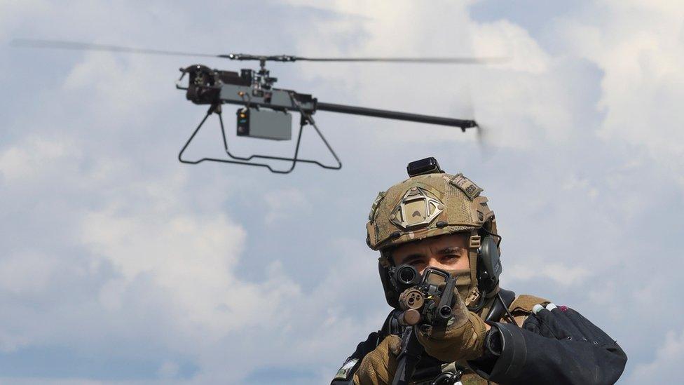 A Ghost drone hovers just behind an armed marine who is pointing his rifle at the camera