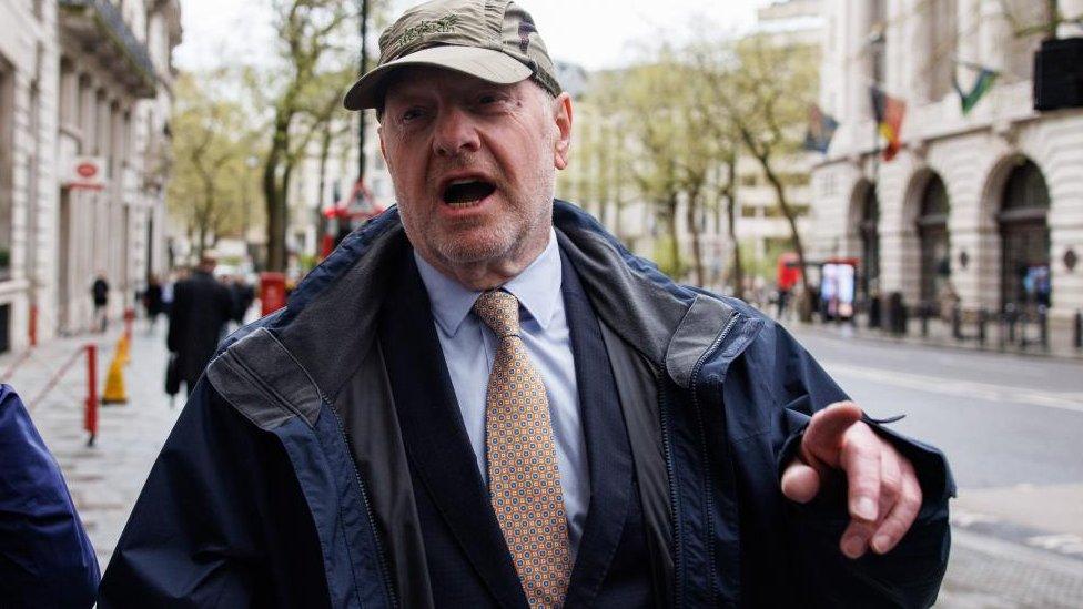 Bates, in a suit, rain jacket and cap, gestures as he speaks