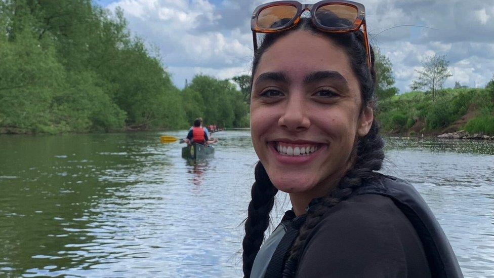 Image of Lauren Bennett. She is pictured sailing on a river, which can be seen in the background. She is looking over her shoulder towards the camera and smiling.