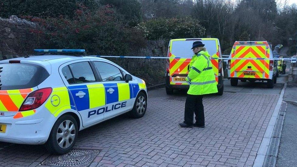 A female police officer wearing a high visibility jacket stands by three police vehicles and a police cordon