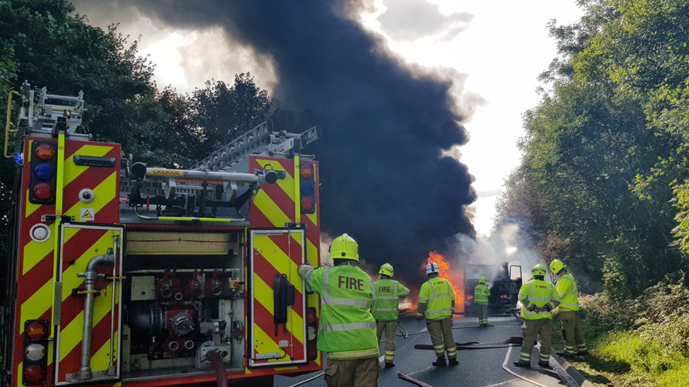 Fire engine with lots of black smoke and flames surrounding a burnt out lorry