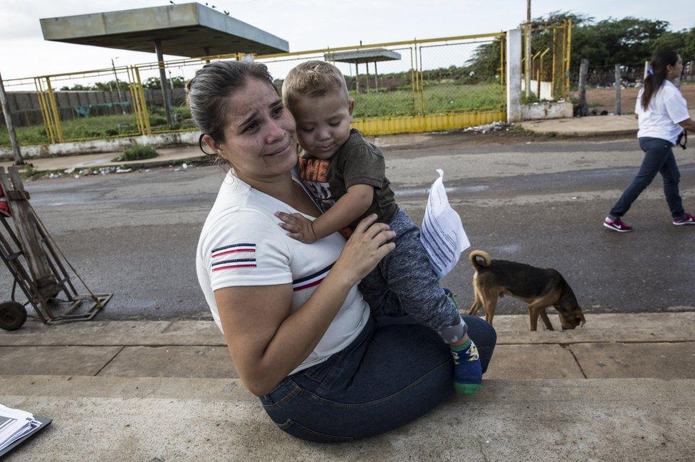 Isobel sits and waits for her husband on the roadside whilst holding her baby.