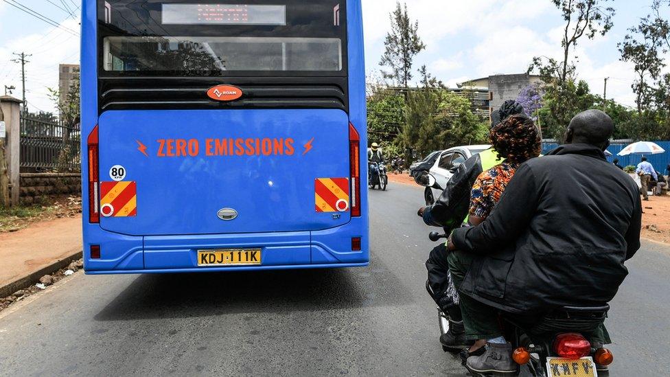 A motorbike rider transports passengers next to an electric bus in Nairobi, Kenya - 2022