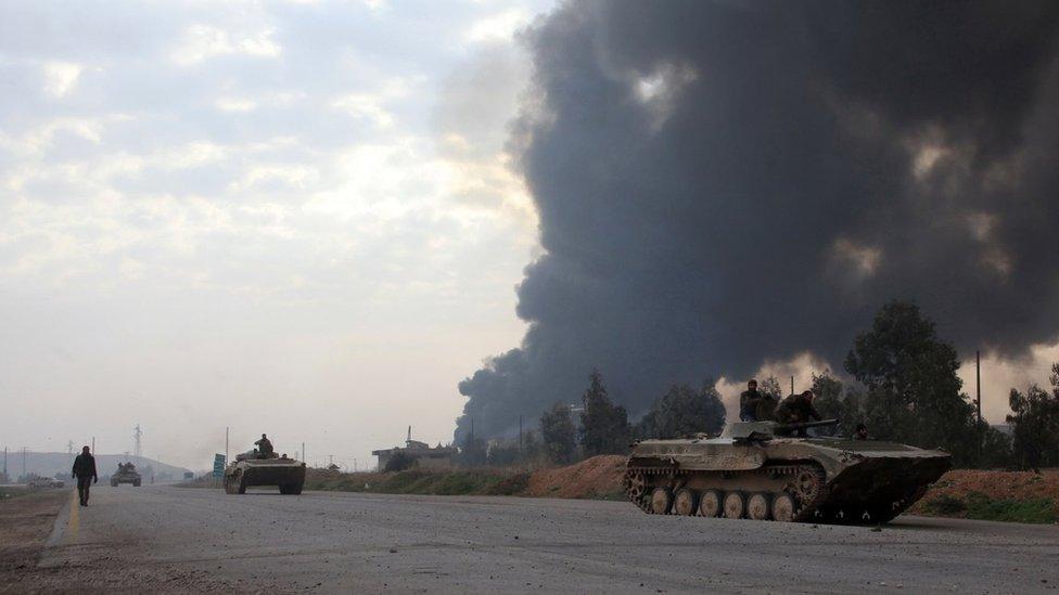 Syrian government forces drive tanks on a road as smoke billows from a power plant outside the northern city of Aleppo (21 February 2016)