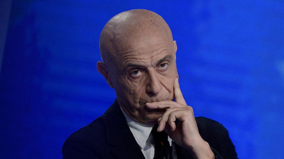 Marco Minniti is pictured against a bright blue background, one hand against his face in a pensive pose