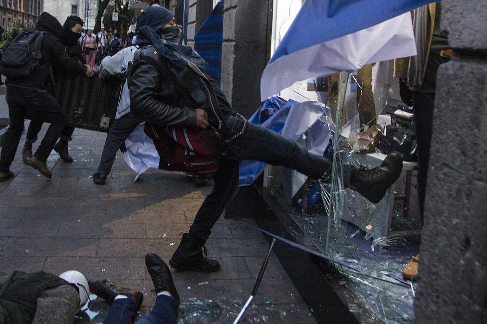 A man in a balaclava smashes a shop window with his boot