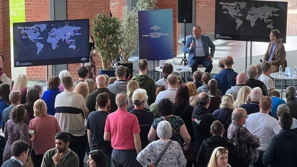 People standing around, chatting, or watching a presentation and discussion with Page Thompson at a public meeting. A screen shows a world map with the words "Why Europe?"