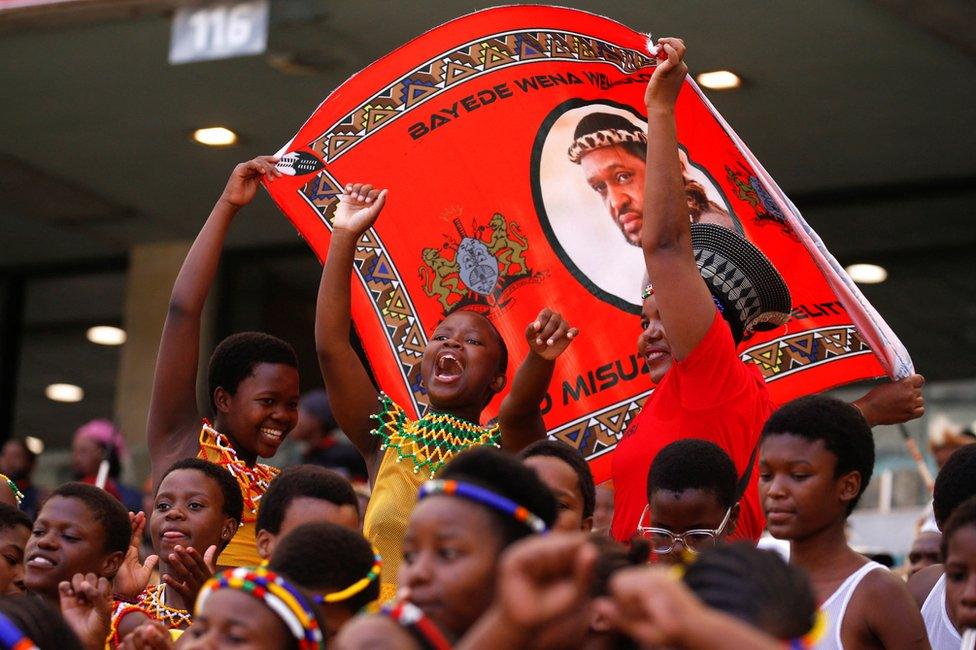 Young people dressed in traditional Zulu attire smile and wave a banner showing King Misuzulu kaZwelithini.