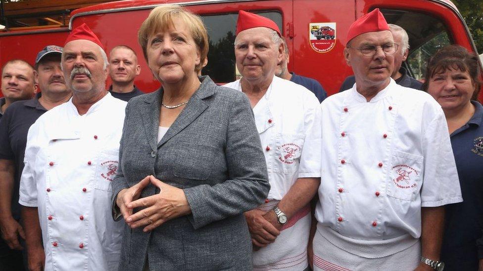 Angela Merkel posing with a group of men in white and red uniforms with red caps, 3 August