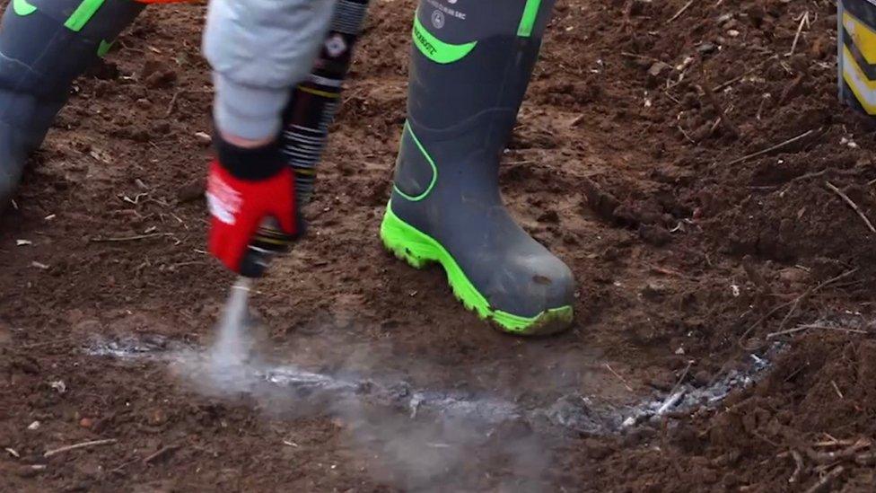 A gloved hand is spraying from a can onto soil. The paint has made a line on the ground. Two wellington boots are also visible.