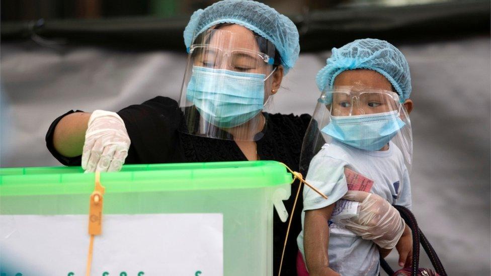 A woman in a mask, hair net and wearing gloves puts her vote in a ballot box while holding her child in Myanmar