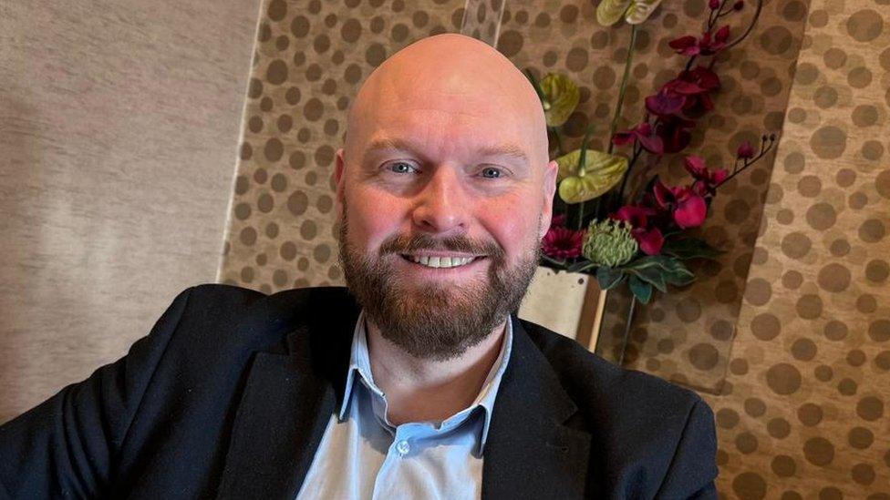 David Jones, a shaven-headed white man in a suit smiles in the camera.