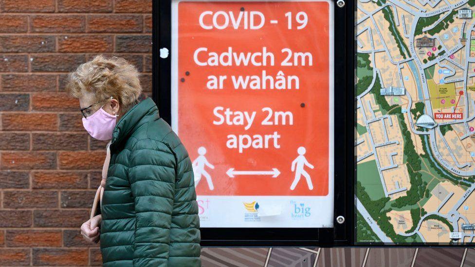 Menyw yn cerdded heibio siop gyda'r arwydd 'cadwch 2m ar wahân'