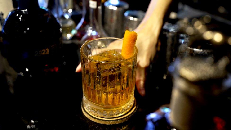 Person places whisky in tumbler on bar with drinks bottles.