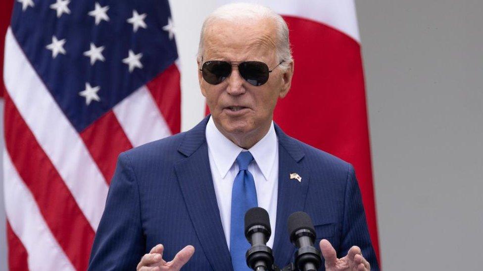 Joe Biden wears a blue suit and black aviator sunglasses. He gestures as he speaking into a microphone