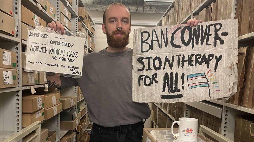 Adam Baker at Norfolk Heritage Centre holding up a poster and a sign that are in the Queer Norfolk archive