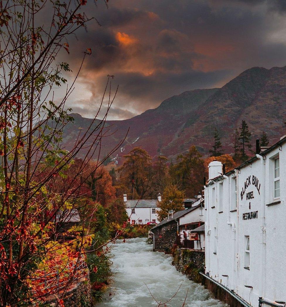 A choppy beck flows by a white-painted hotel with large hills and orange and black clouds beyond