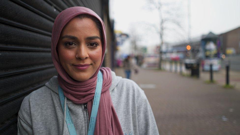 Hana, a young woman wearing a hijab and lanyard, outside a shop on a road in Leeds