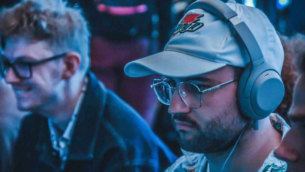 A man wearing a baseball cap and headphones looks at a screen while playing a video game. He's wearing glasses and a white shirt with a floral pattern on it. The band of his grey over-ear headphones is over the top of his grey baseball cap, which has a Gatorade logo - a red lightning bolt with green lettering through the middle. He's concentrating hard.