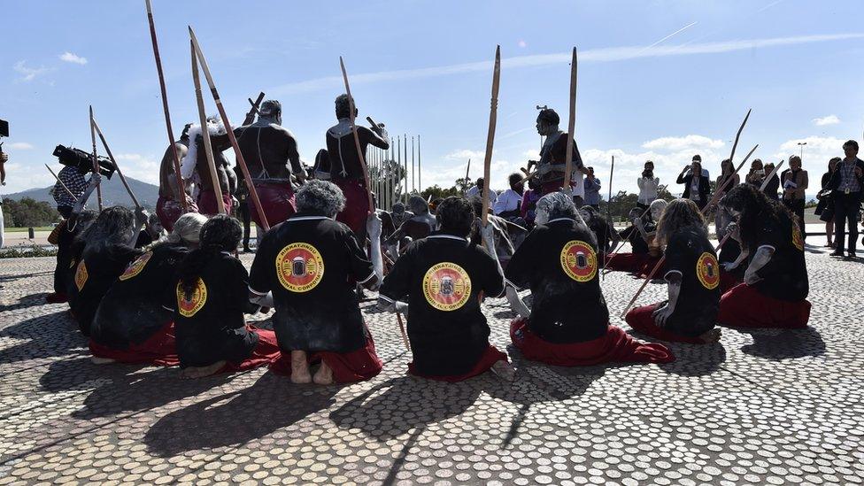 A group of dancers kneel in a circle as a smaller group sand up in the centre