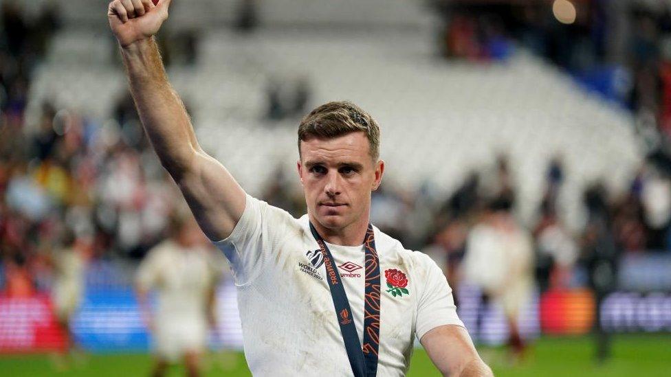 Man with short light-coloured hair has one arm in the air at a rugby ground