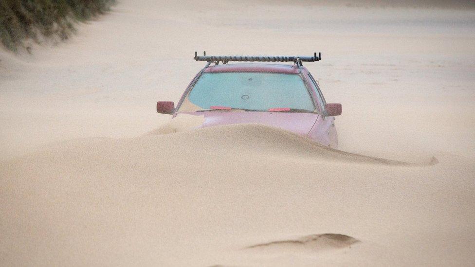 Car part-buried in sand on Fistral Beach, Cornwall