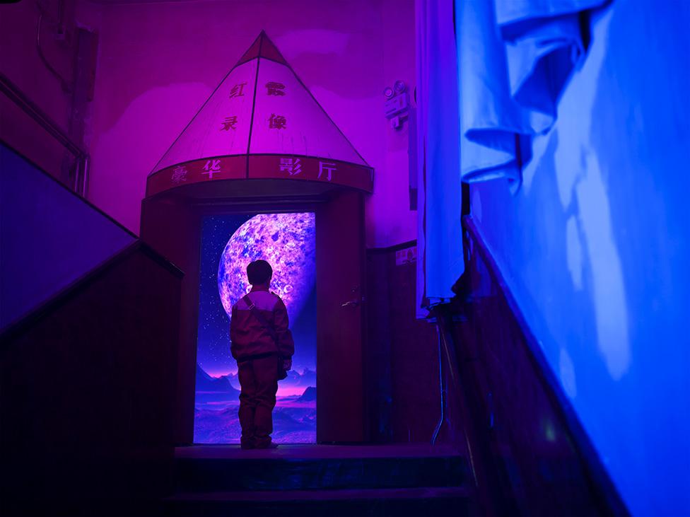 A photo by Cao Fei showing a boy looking at a planet through a doorway in the shape of a rocket