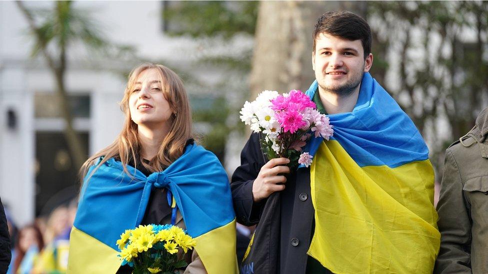 Two people wrapped in Ukraine flags carry flowers