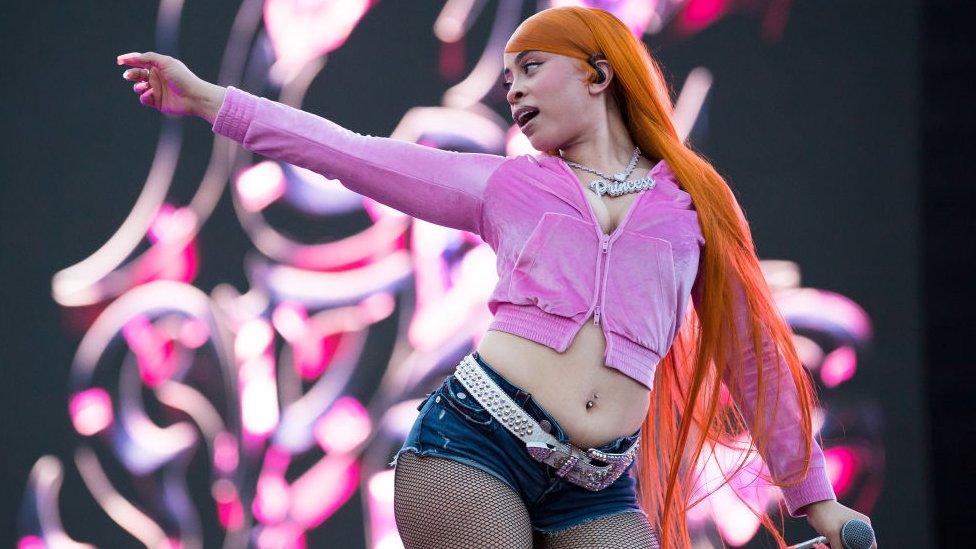 American rapper Ice Spice performs onstage during day one of Rolling Loud Miami at Hard Rock Stadium on July 21, 2023 in Miami. Ice Spice wears a long orange wig down past her back, a cropped pink velvet bomber jacket and denim shorts over fish net tights, accessorised with a white studded belt, diamond necklace reading 'Princess' and a belly button ring. She holds a microphone with her right hand and points off stage with her left, following the direction with her face.