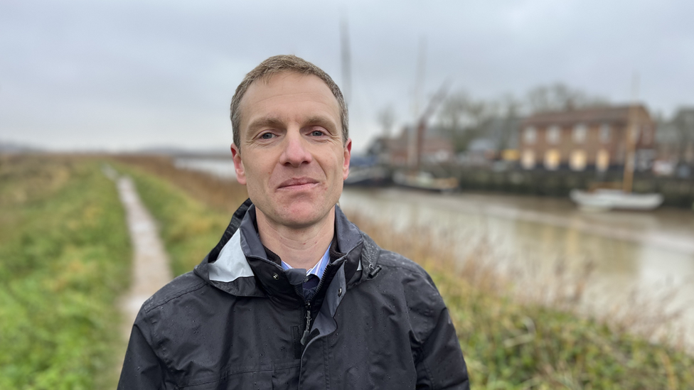 Man with short light-brown hair wearing a blue raincoat stands alongside the estuary