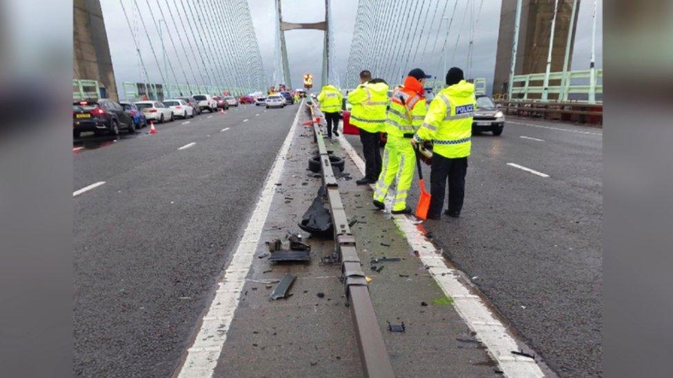 Image of the M4 Prince of Wales Bridge. Workers wearing high vis jackets can be seen stood by the damaged central reservation. Lane 3 on either side of the reservation is closed and cars can be seen queuing in the other two lanes.