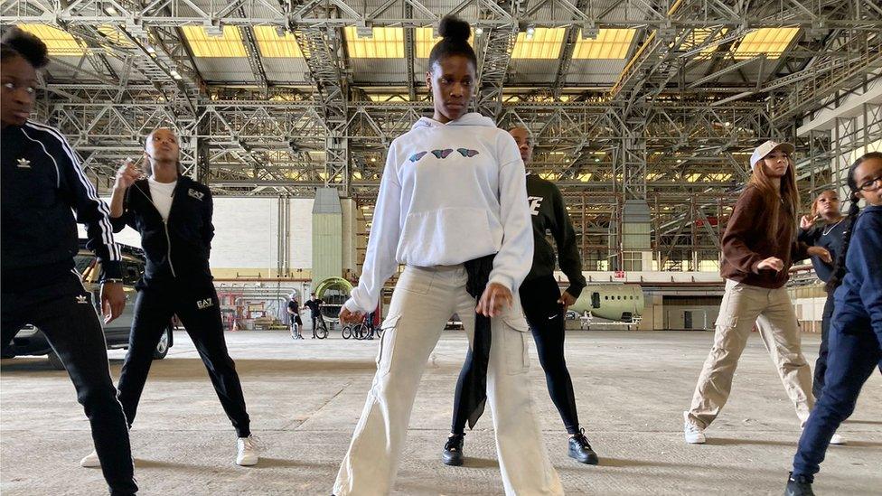 Dancers practicing inside a hangar. 7 dancers are pictured