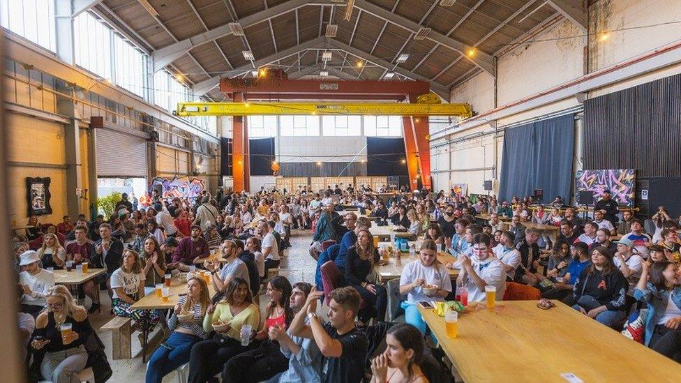 Propyard venue with people sitting at tables and chairs watching a big screen