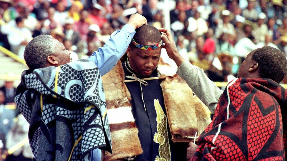 Two traditional chiefs put a calfskin headband and feather on the new monarch's head