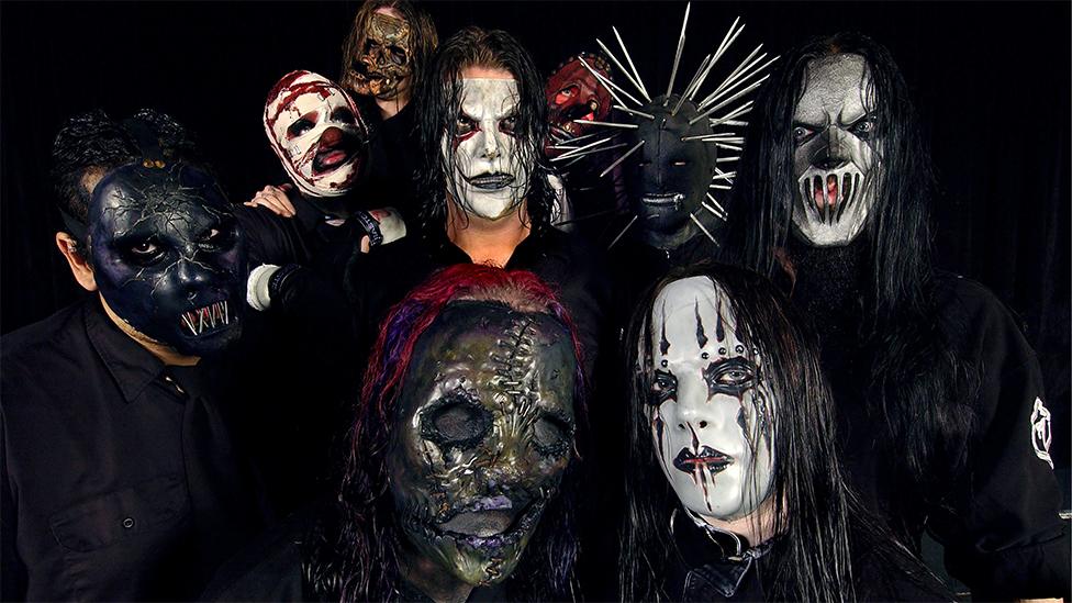 Eight members of Slipknot, wearing a mix of dark skull masks and white horror style masks, all while looking at the camera.