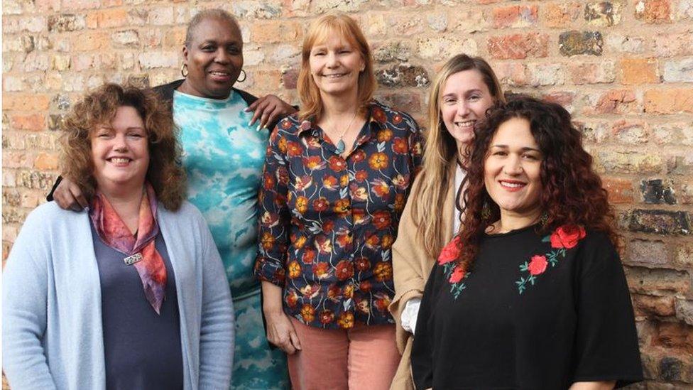 Five women stand smiling in front of a brick wall