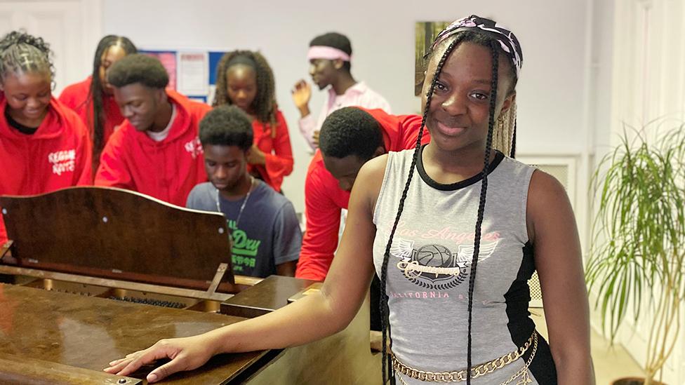 Stephanie Horacio from the Reggae Roots choir looking at the camera, wearing braids and a grey top, with her fellow choir members in the background.