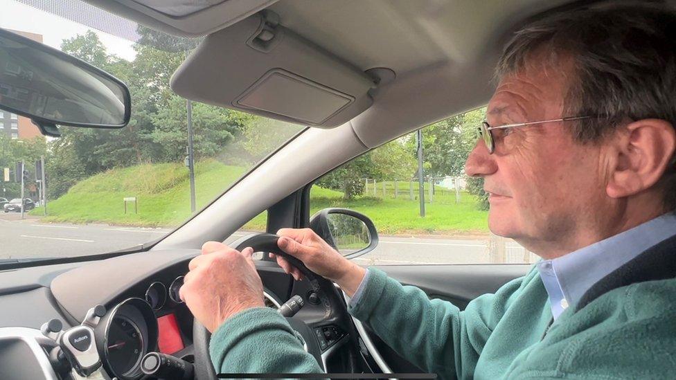 A man in a green jumper with glasses on behind the wheels of a car