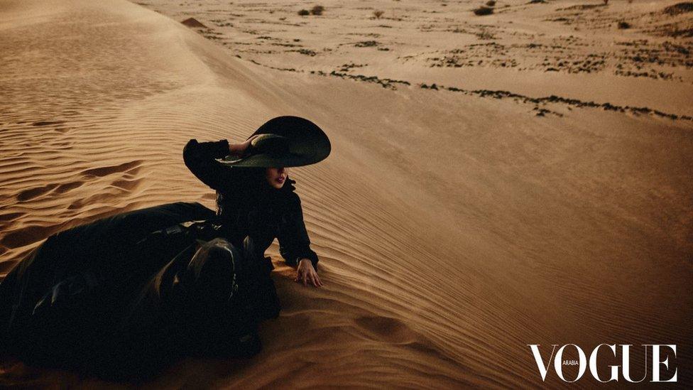 Princess Hayfa Bint Abdullah Al Saud, in a large black hat and dress, lying on a sand dune