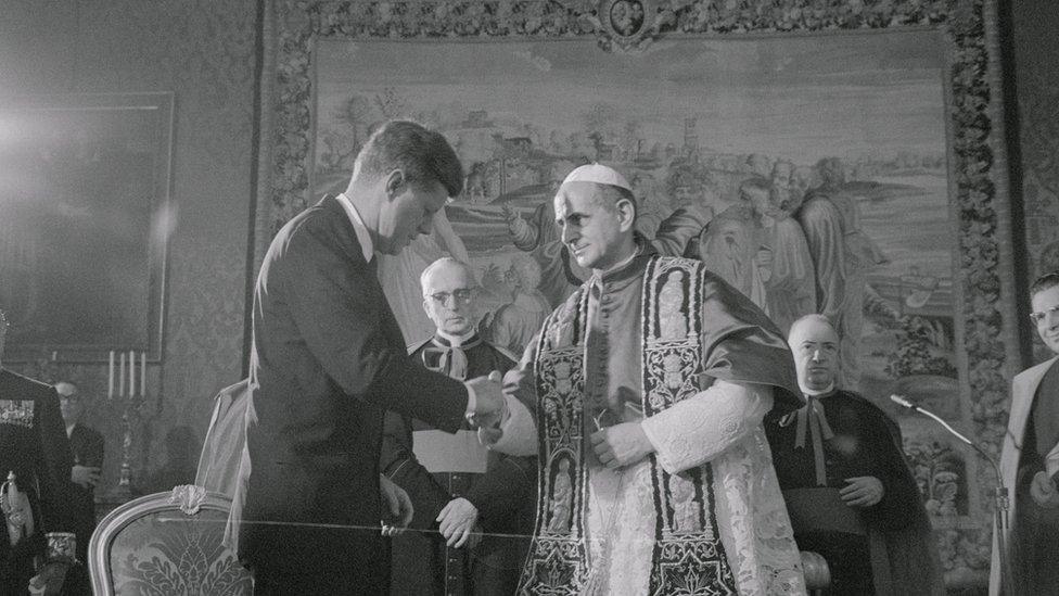 A black and white photograph, 1963, shows JFK shaking the hand of the pope, flanked by multiple high-ranking clergy amid opulent surroundings in the Vatican