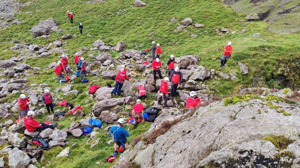 A large group of volunteers in red jackets and white helmets prepare ropes