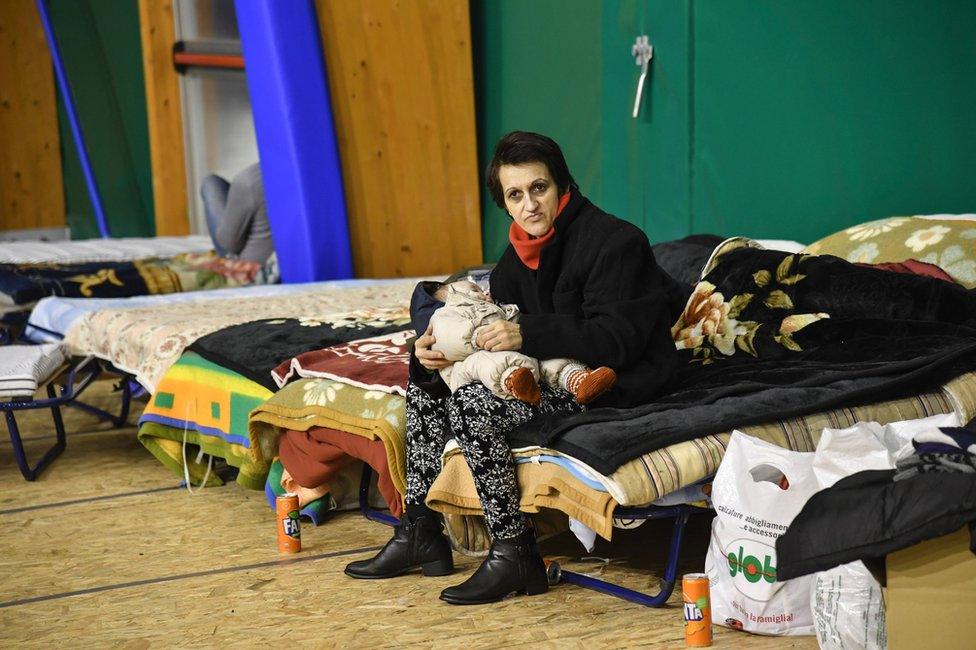 A woman holding a child in her arms sits on a bed as residents from the village of Montereale set up for the night in a large tent on a nearby sports ground in Montereale, Abruzzo, 18 January