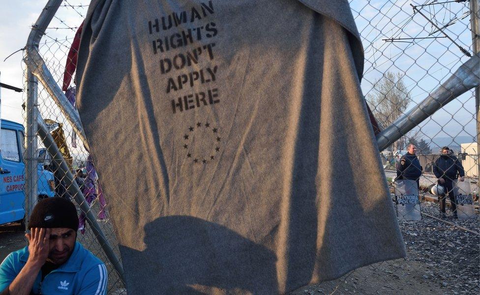 Migrant sits next to a blanket displaying message "Human rights don't apply here" at Idomeni