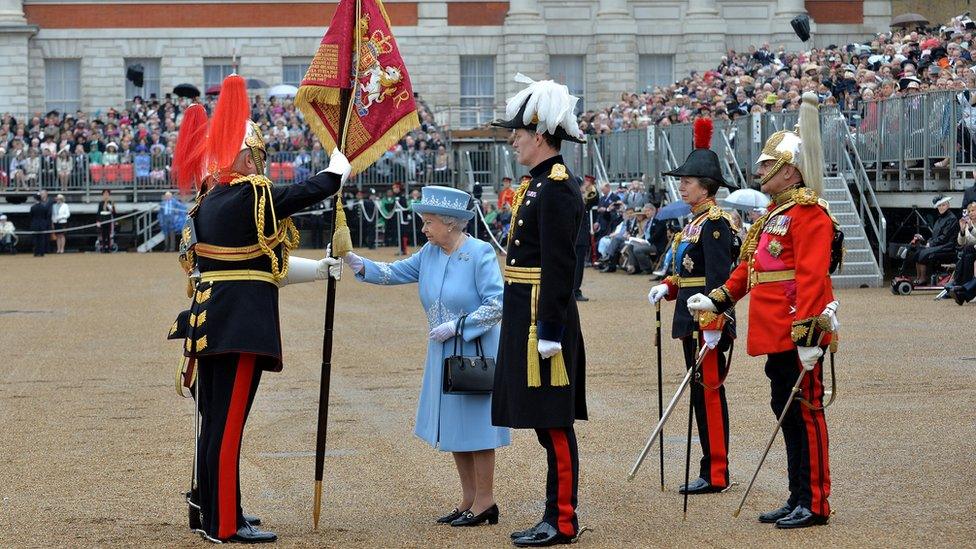 The Queen, wearing a pale blue coat and hat, presenting a flag to a soldier wearing a black uniform with gold braids and a helmet with a red plume, in front of a crowd of seated spectators.