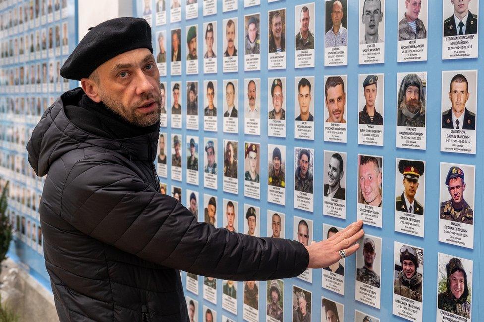 Father Dmitriev stands at the portrait wall for those who died, pointing to the first man from the priest's brigade to lose his life in the war.